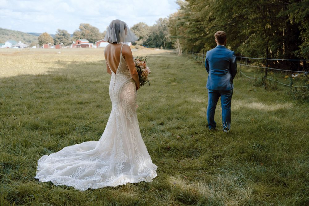 A couple wearing wedding attire stands apart in a grassy meadow with a long flowing lace dress train visible.