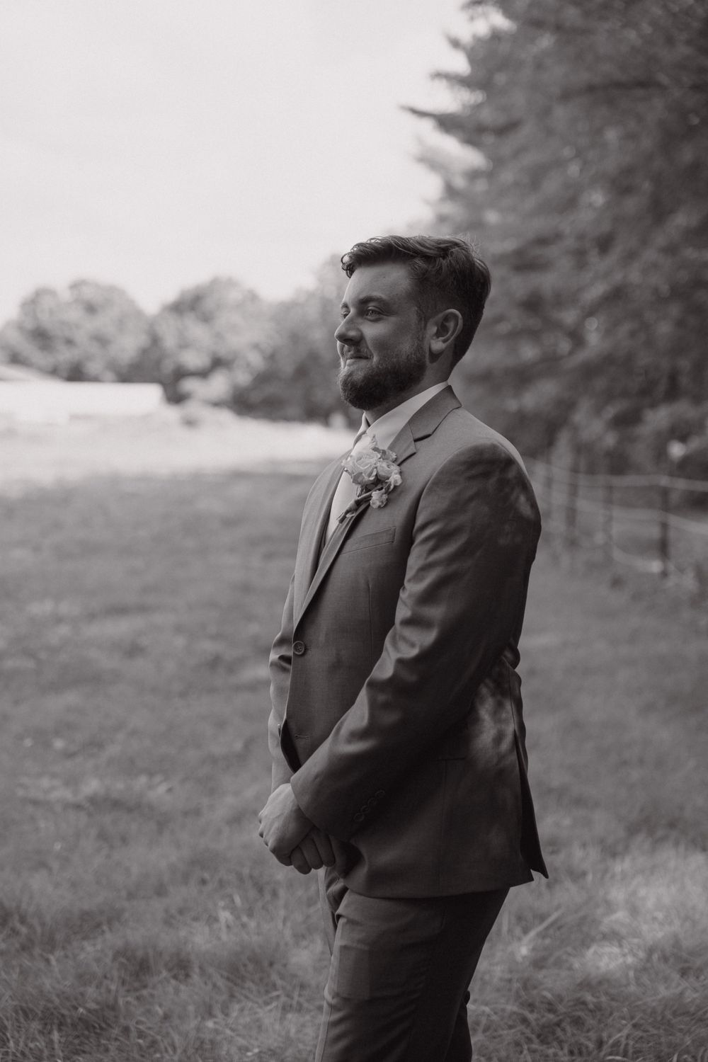 A groom in a gray suit stands alone outdoors during a wedding day photoshoot.