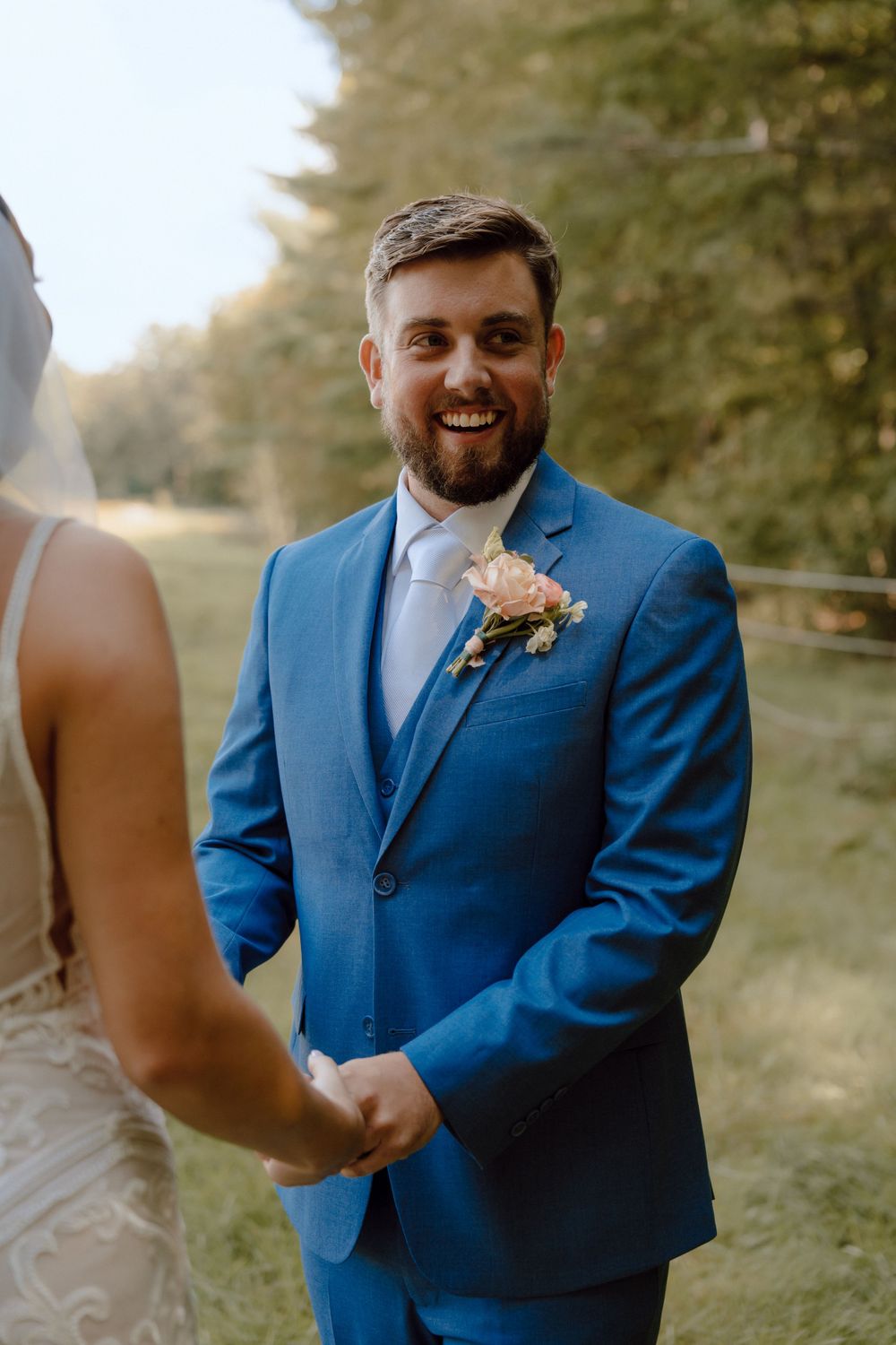 A groom in a bright blue suit and blush boutonniere smiles warmly during a wedding ceremony outdoors.
