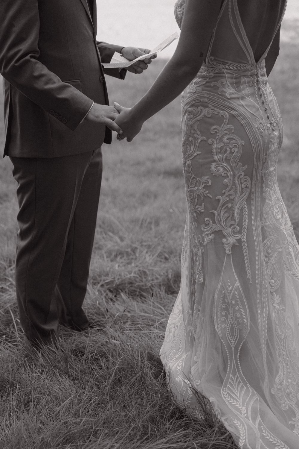 A close-up black and white photo of a bride in an intricate lace dress holding hands with her groom during a ceremony.