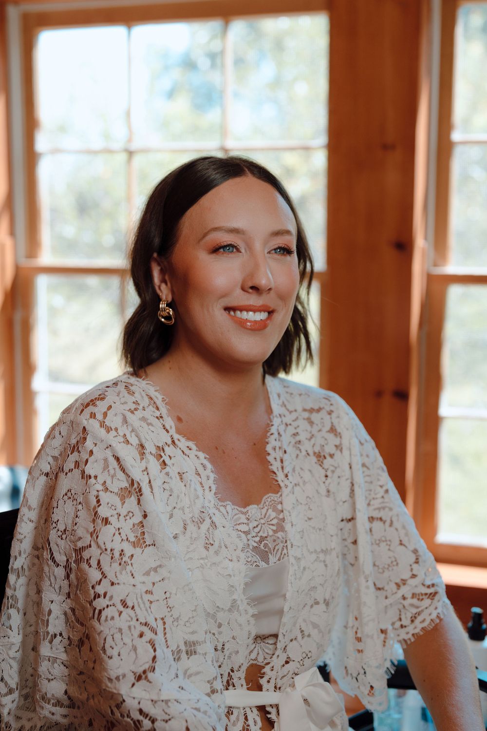 A smiling woman wearing a delicate white lace overlay sits near sunlit windows.