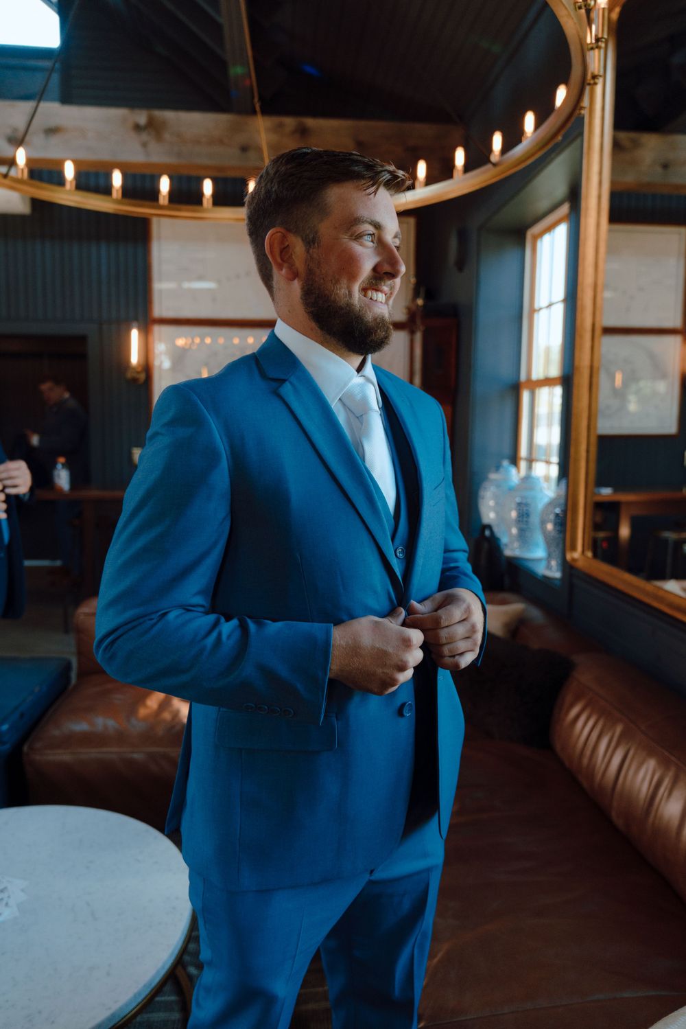 A groom in a vibrant blue suit adjusts his jacket while standing in an elegant room with circular lighting overhead.