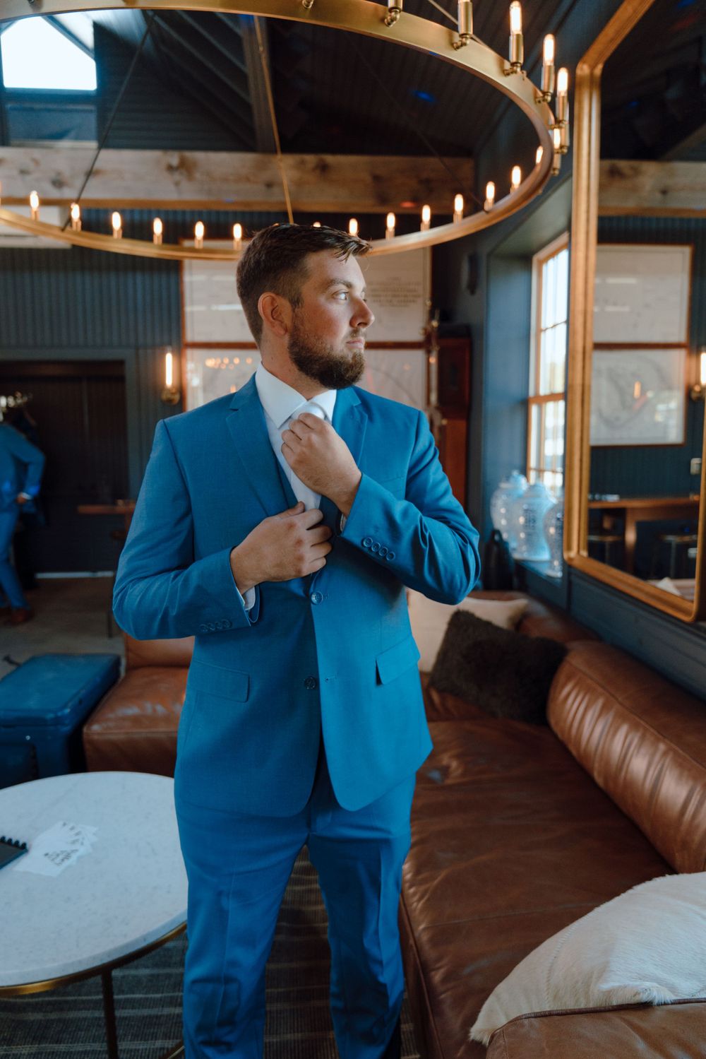 A groom in a bright blue suit adjusts his tie while standing in an elegantly lit venue.