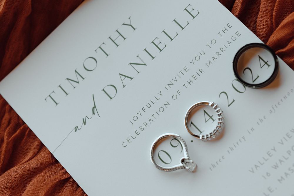 Wedding invitation card with elegant white engraving laid on a rust colored fabric with engagement and wedding rings beside it.