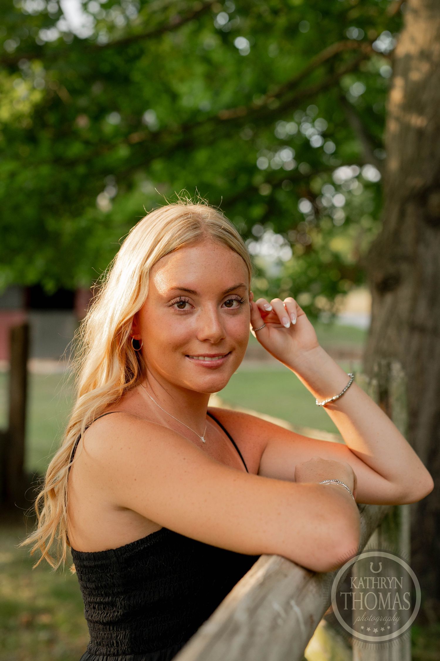 A woman in a black top poses against a natural backdrop of green trees during sunset.
