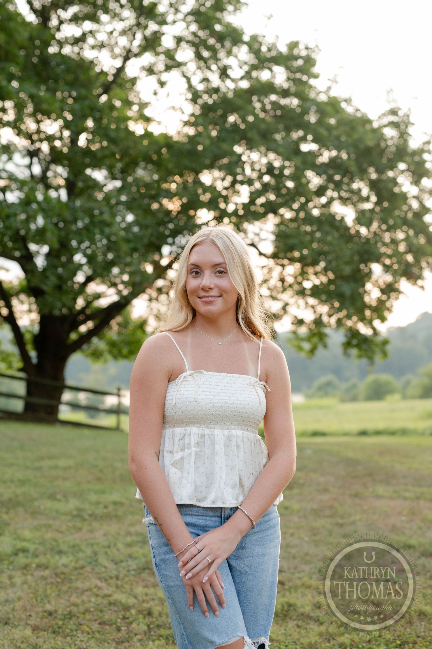 Outdoor summer portrait in white top and jeans standing in field with trees at sunset.