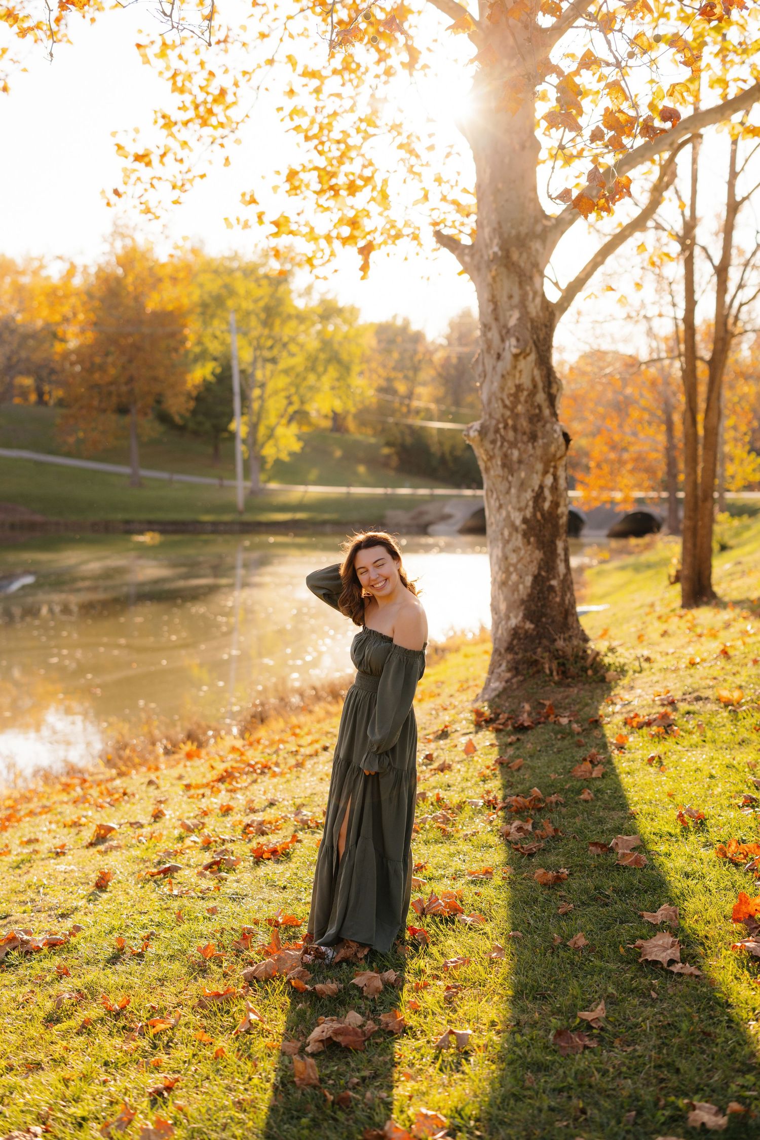 Person in a long green dress poses by a tree during golden hour autumn sunset near a pond with fallen leaves.