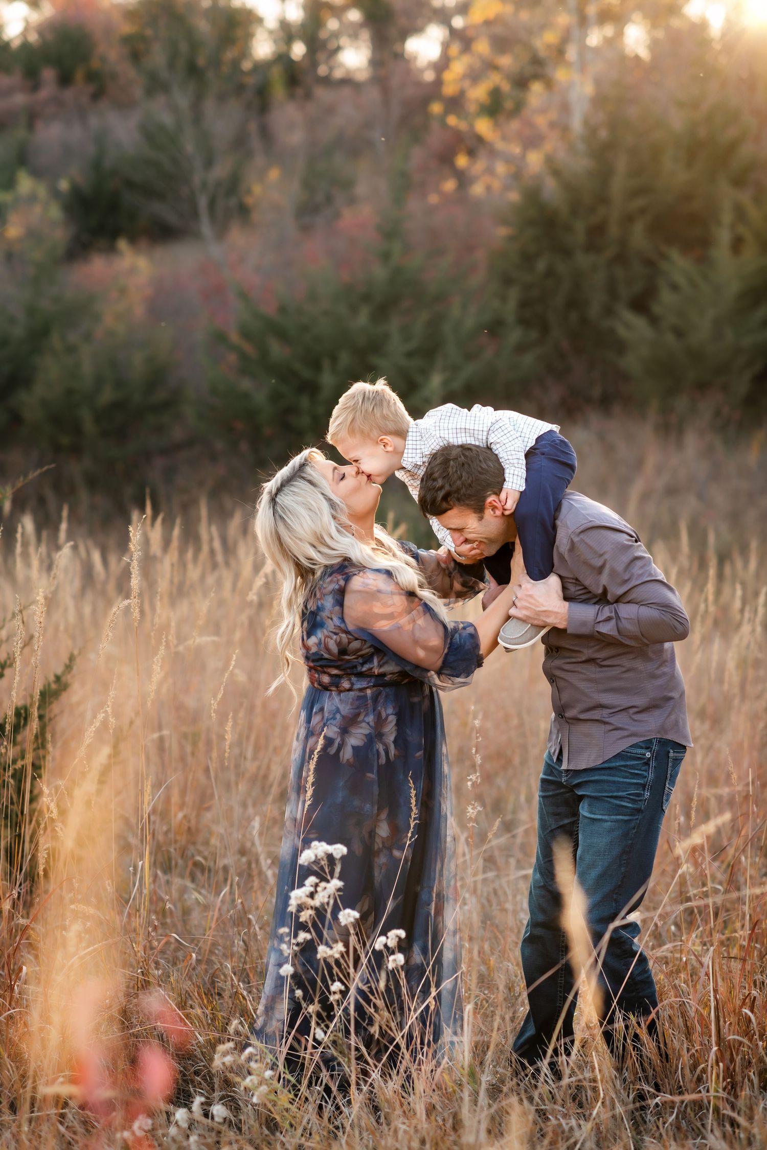 A couple shares an intimate moment in a golden field at sunset among tall grass and evergreen trees in the background.