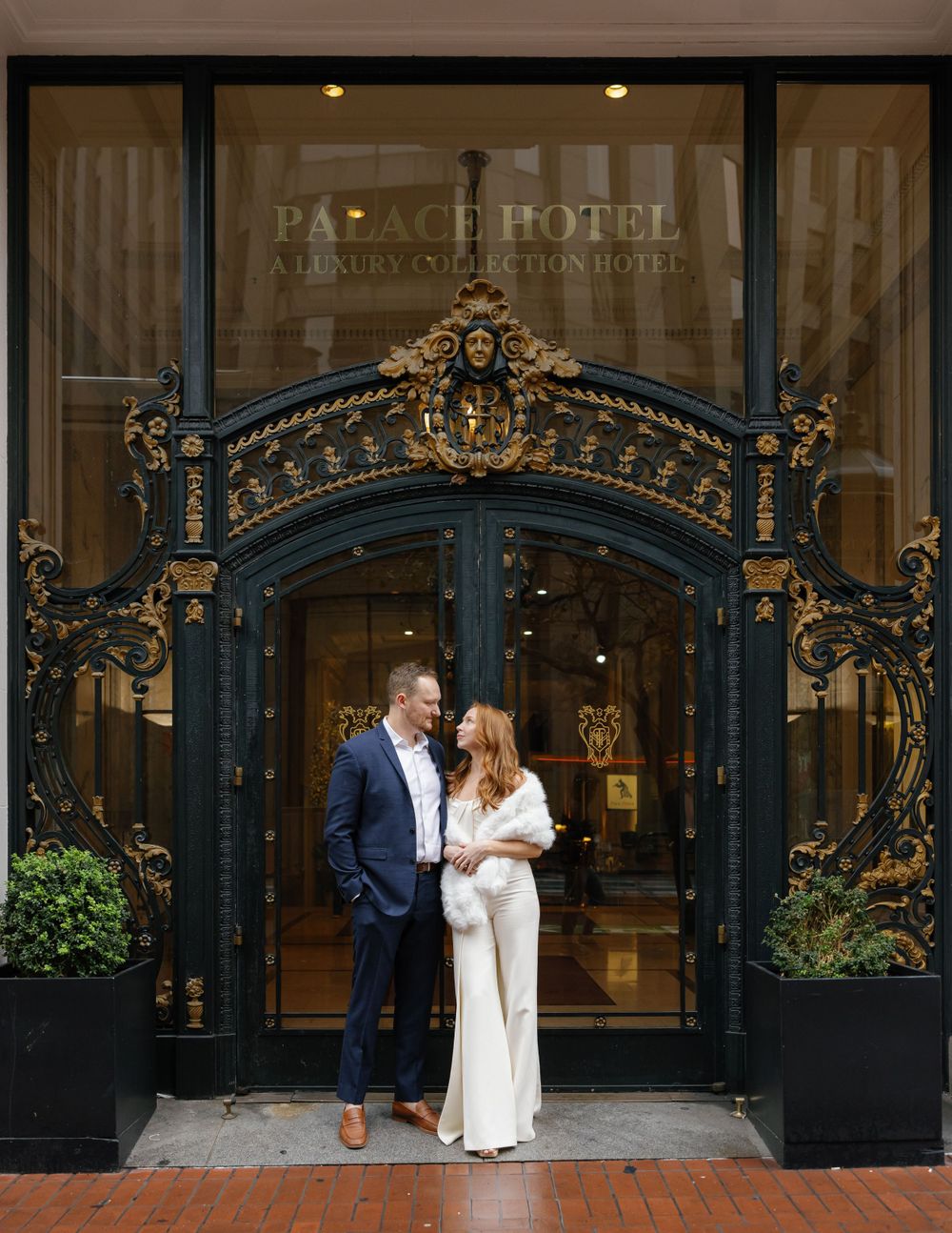 Elegant couple posing in front of ornate Palace Hotel entrance with decorative black and gold archway in San Francisco.