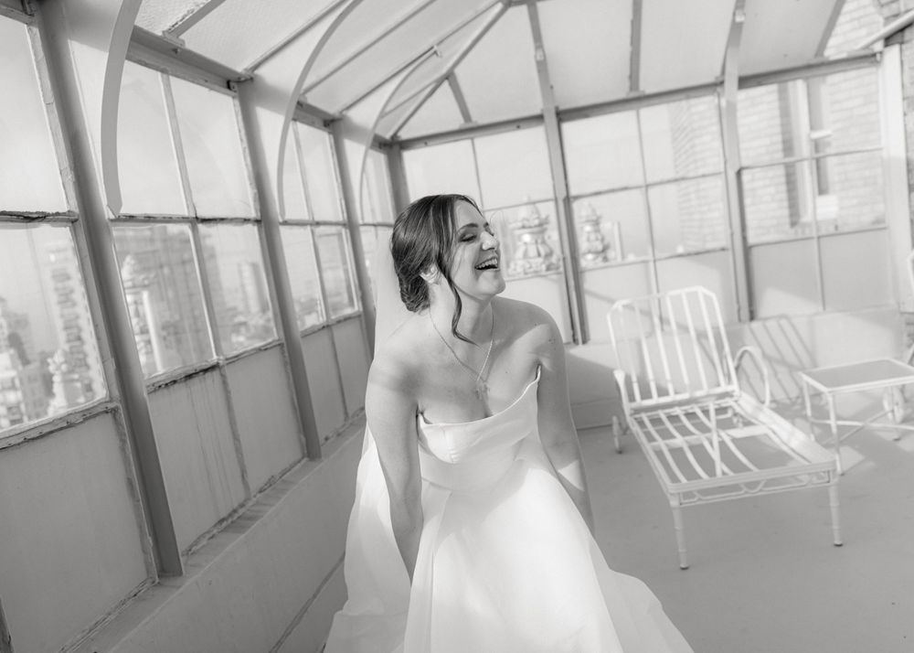 A person in a white wedding dress laughs joyfully while standing in a bright sunroom with white chairs.