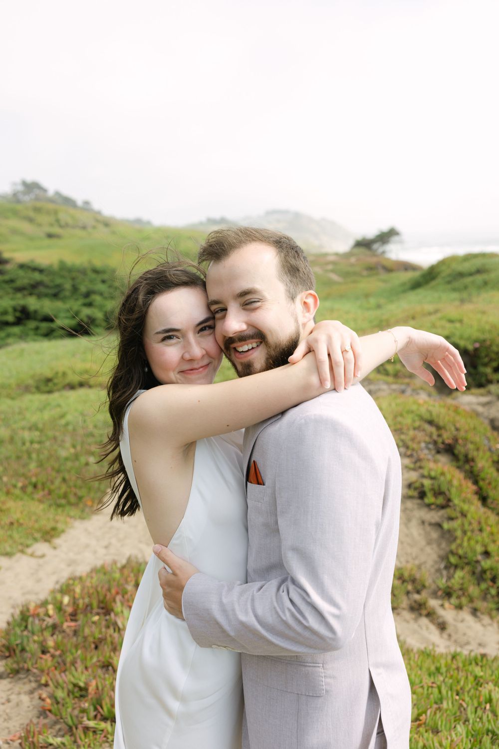 A romantic couple in light-colored wedding attire embrace on a scenic hillside with green grass and mountains in the distance.