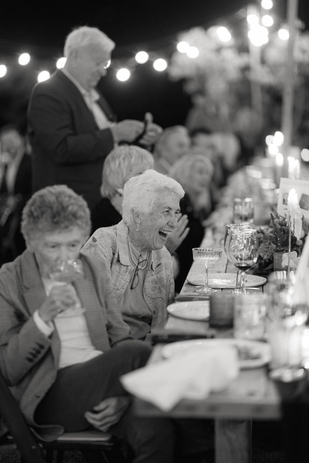 A candid black and white photograph of wedding guests laughing and enjoying themselves at an elegant candlelit dinner table.