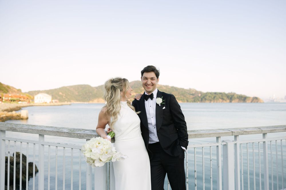 A bride and groom share a romantic moment on a balcony overlooking a scenic coastal bay at sunset.