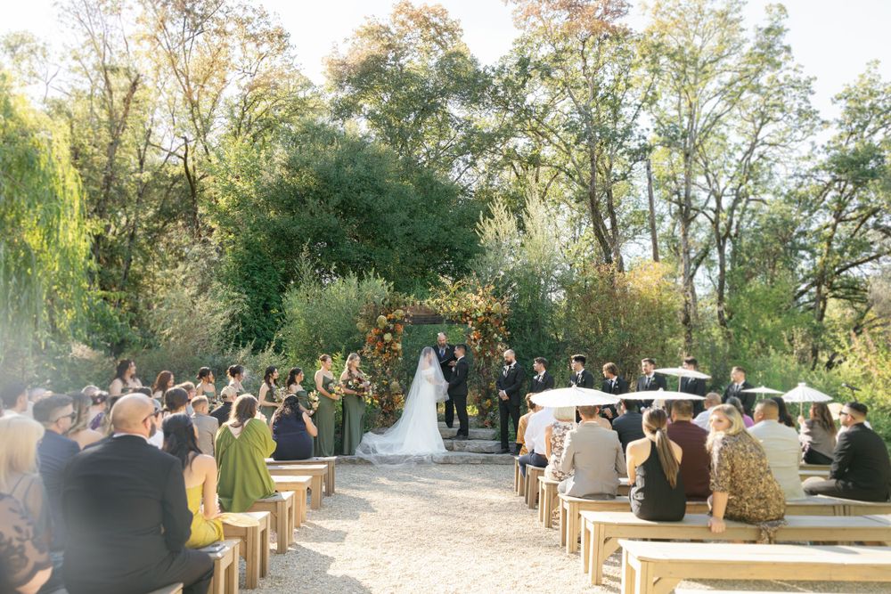 Outdoor wedding ceremony taking place on a garden path with guests seated on wooden chairs amid autumn trees.