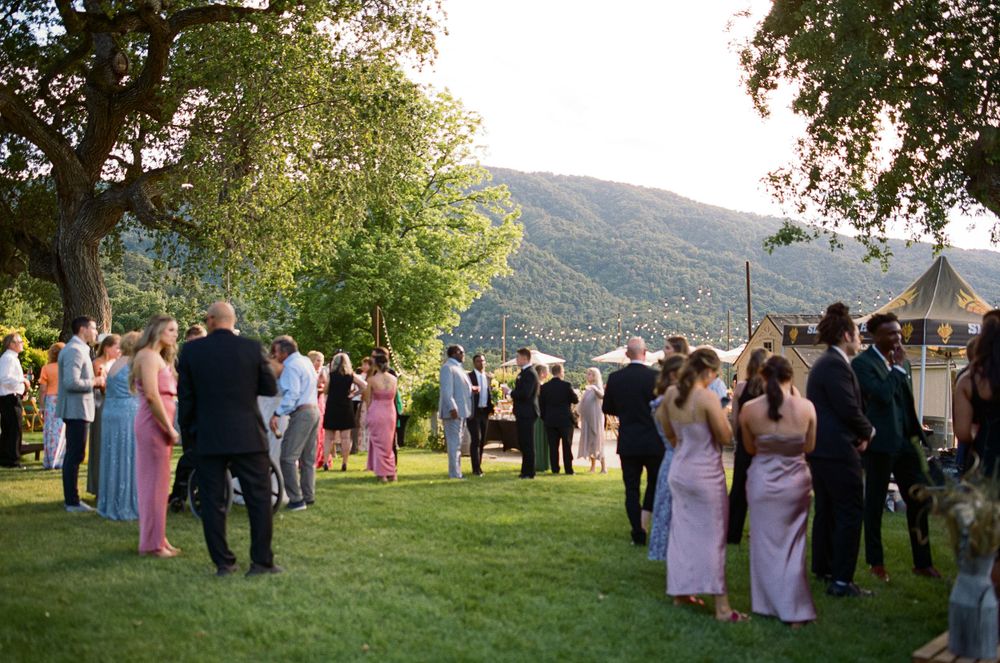 Wedding guests gather on a lush green lawn at an outdoor celebration with mountain views in the background.