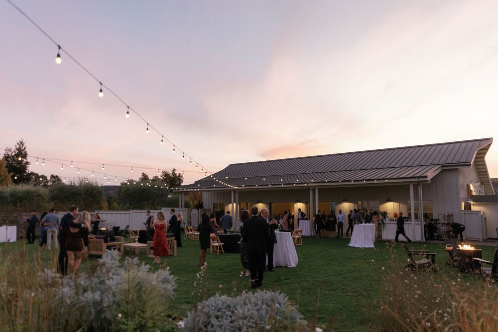 Outdoor evening wedding reception with string lights and rustic barn venue at sunset.