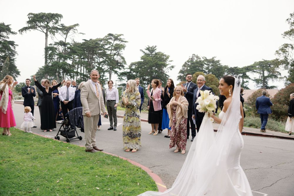 Newlyweds and wedding guests gather on a paved pathway surrounded by lush green trees at an outdoor ceremony.