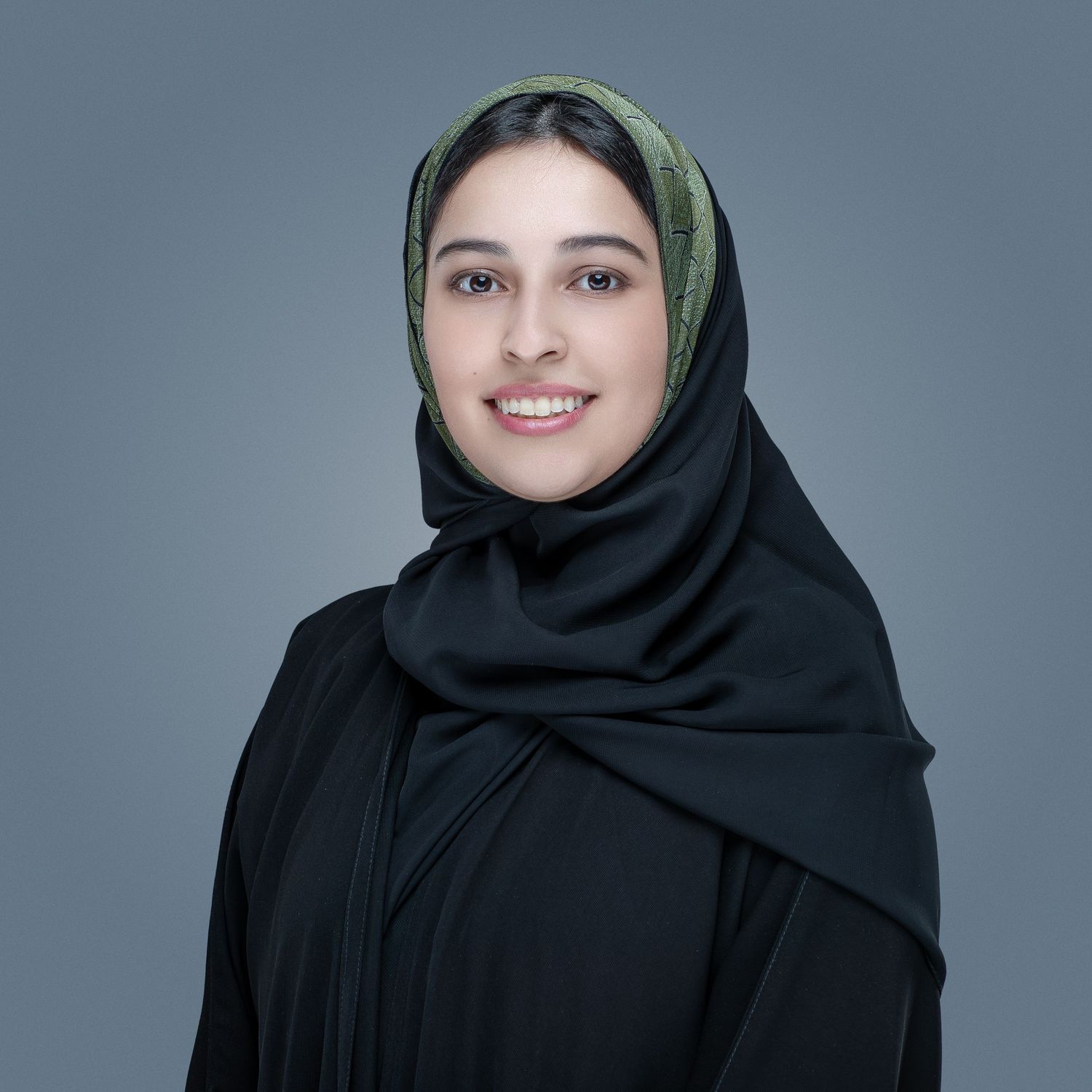 Portrait of someone wearing a black hijab with an olive green accent against a gray background, smiling warmly.