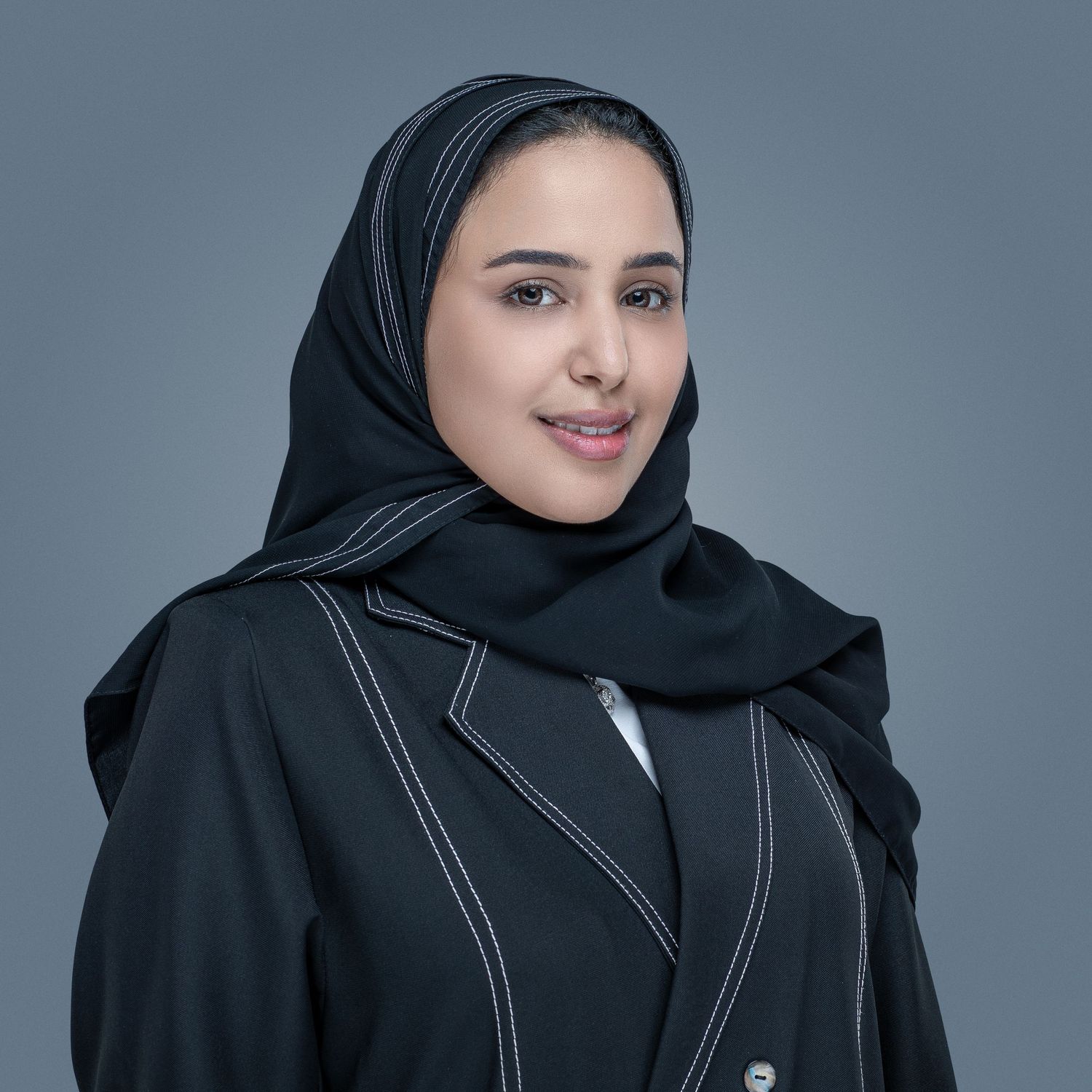 Professional studio portrait of a person wearing a black hijab and blazer with contrast stitching against a gray background.