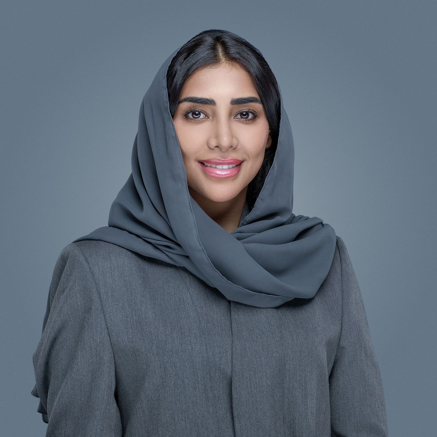 Woman wearing a gray hijab and matching sweater poses against blue-gray background with gentle smile.
