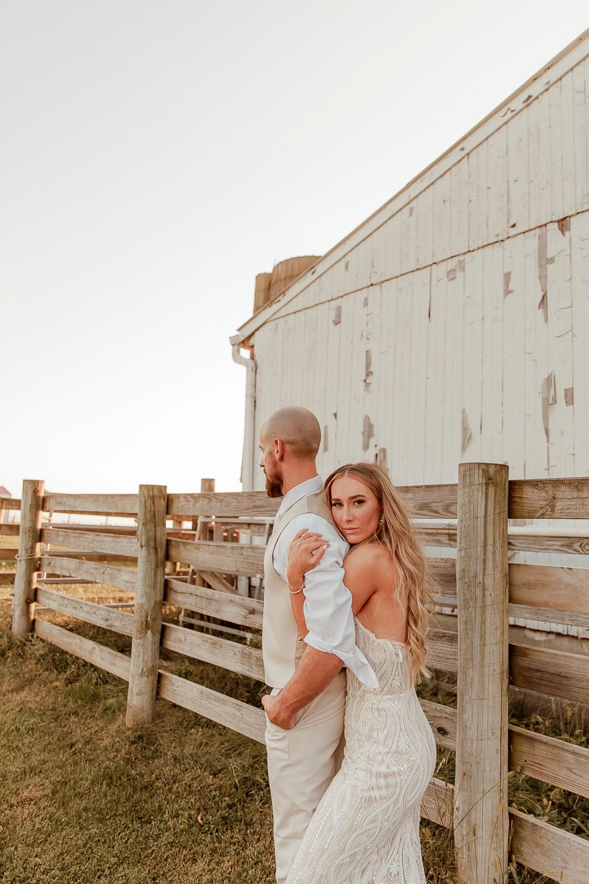 A couple poses together by a rustic white barn and wooden fence during a sunset wedding photoshoot.