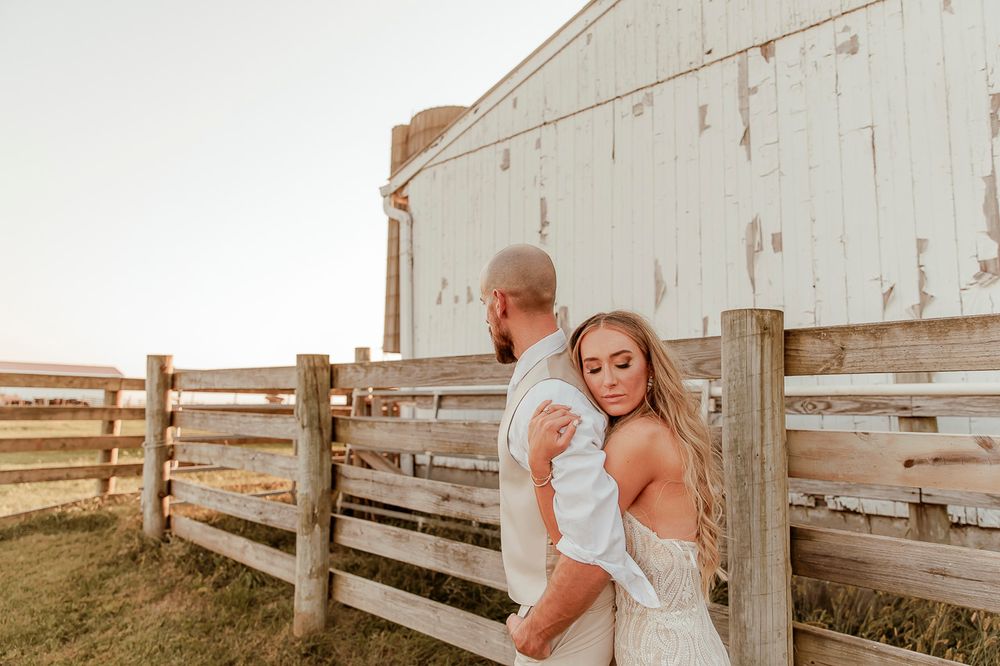 A romantic sunset photo of a couple posed against a rustic wooden fence and white barn in warm golden light.