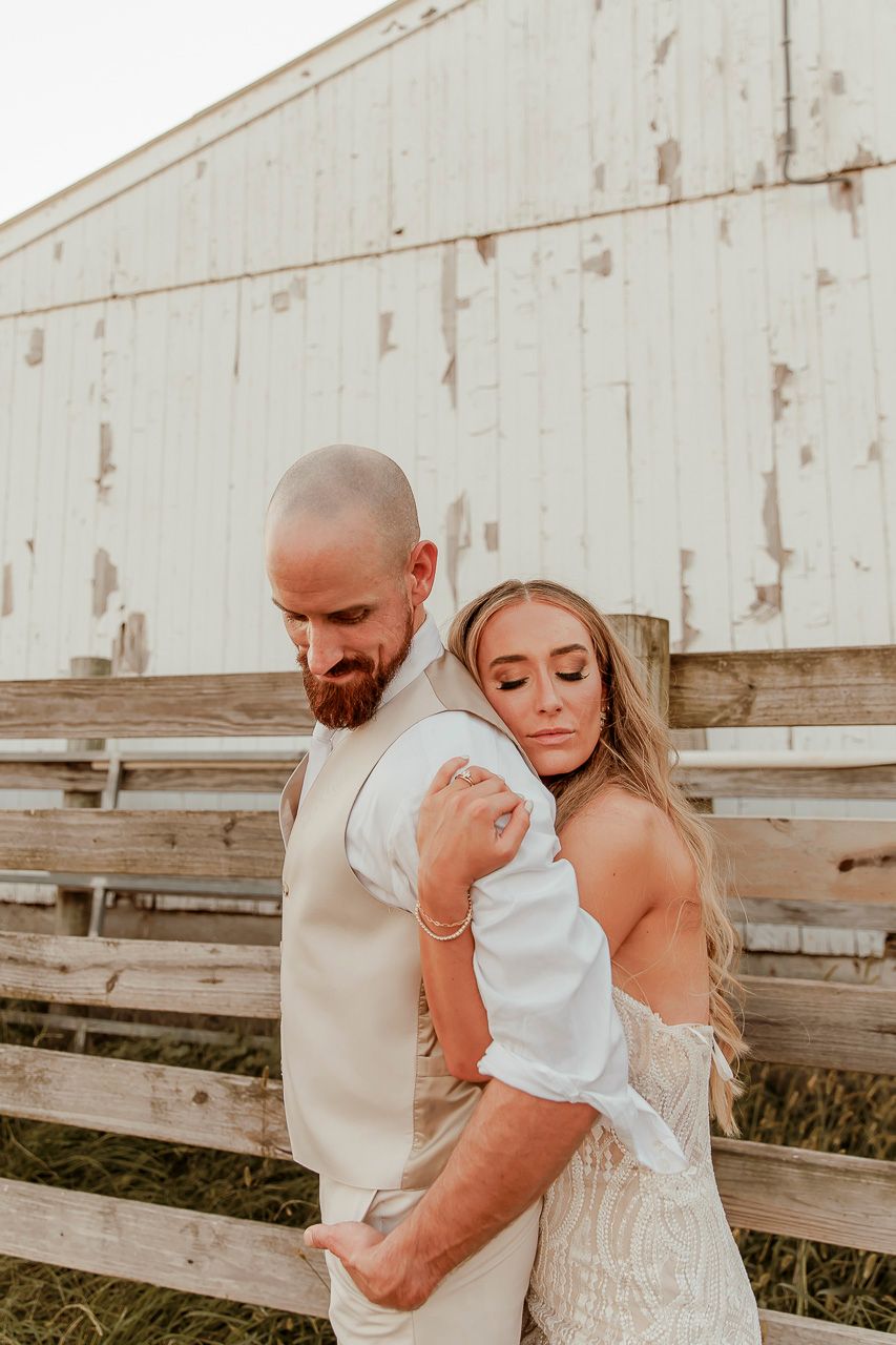 A romantic rustic wedding portrait of a couple embracing by a white barn at sunset.