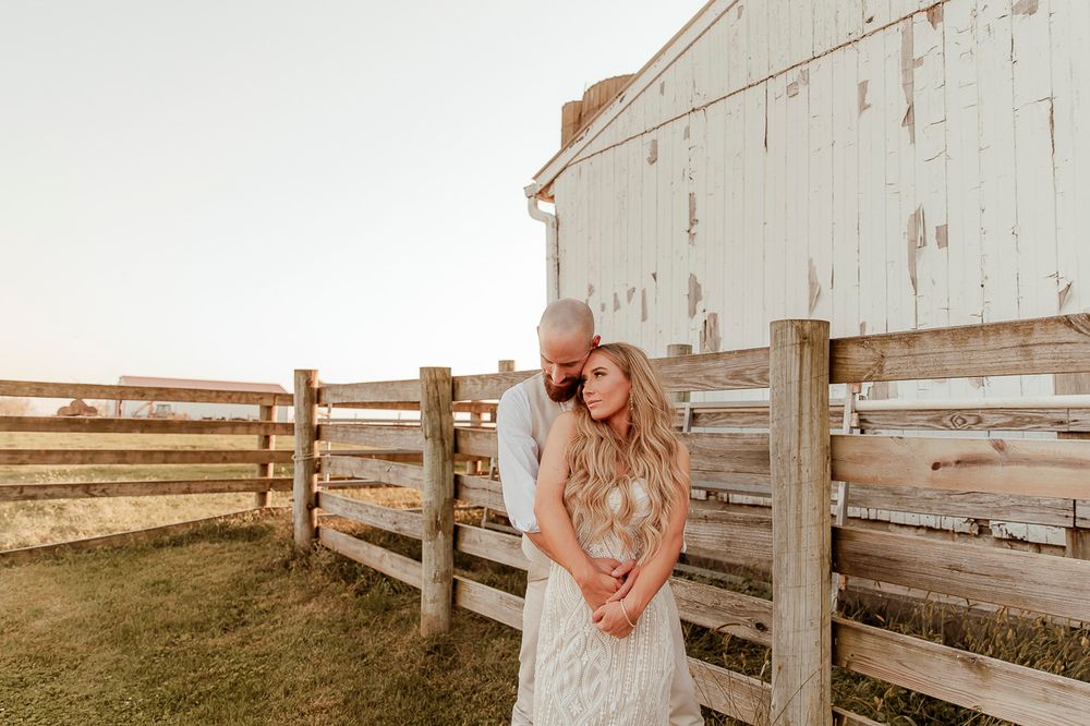 Rustic country sunset scene with couple embracing by weathered white barn and wooden fence.