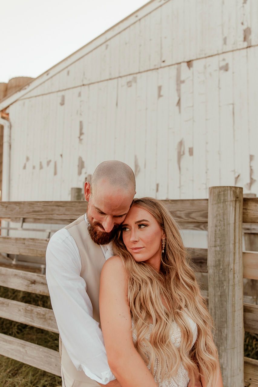 A romantic couple embraces in front of a rustic white barn during a golden hour wedding photoshoot.