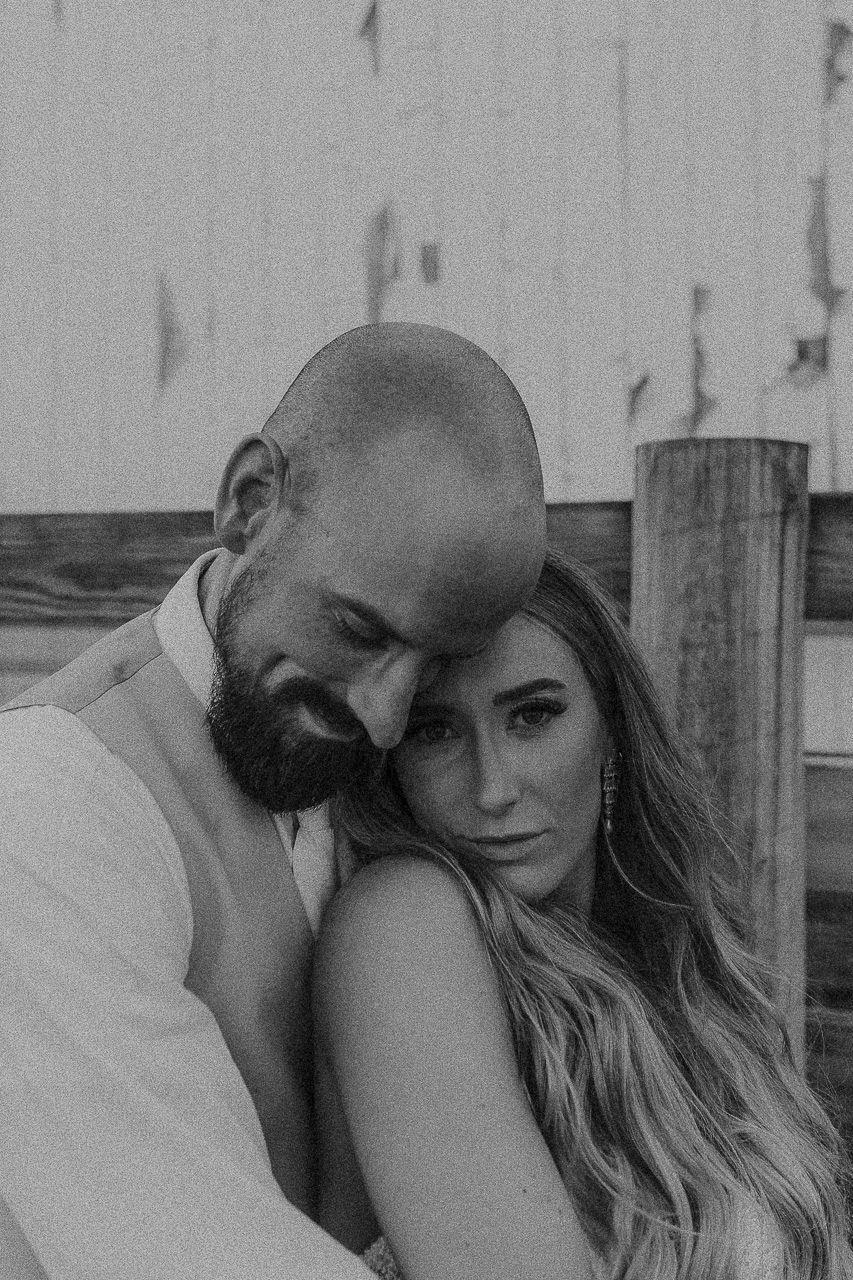 Intimate black and white portrait of a couple embracing outdoors by wooden posts at dusk.