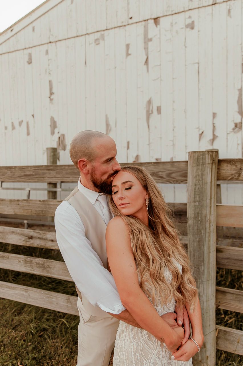 Romantic couple embrace in front of rustic white barn and wooden fence at sunset.
