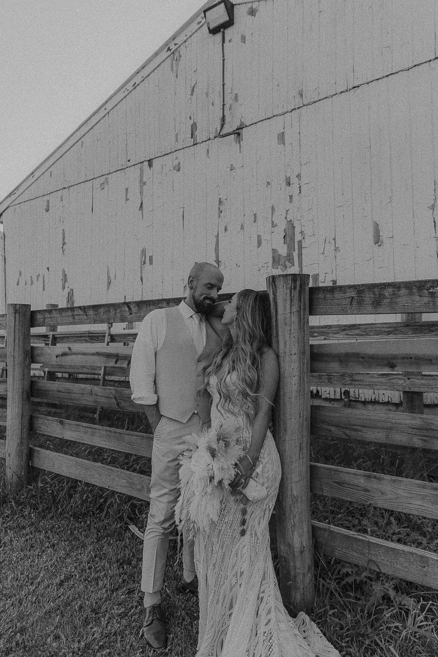 Black and white rustic wedding photo of a couple embracing by a wooden barn fence.