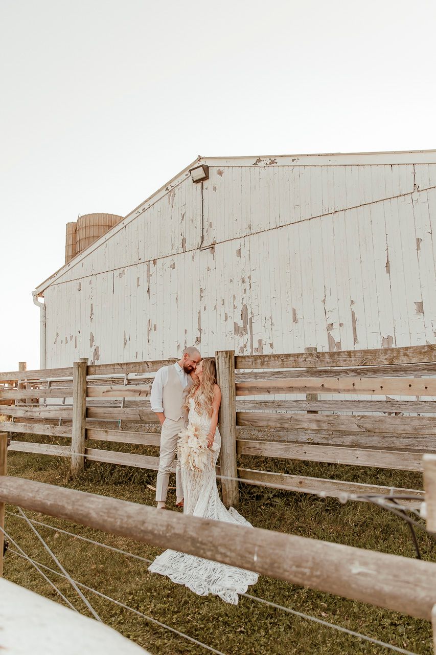 A romantic couple poses against a rustic white barn and wooden fence during golden hour on a farm.