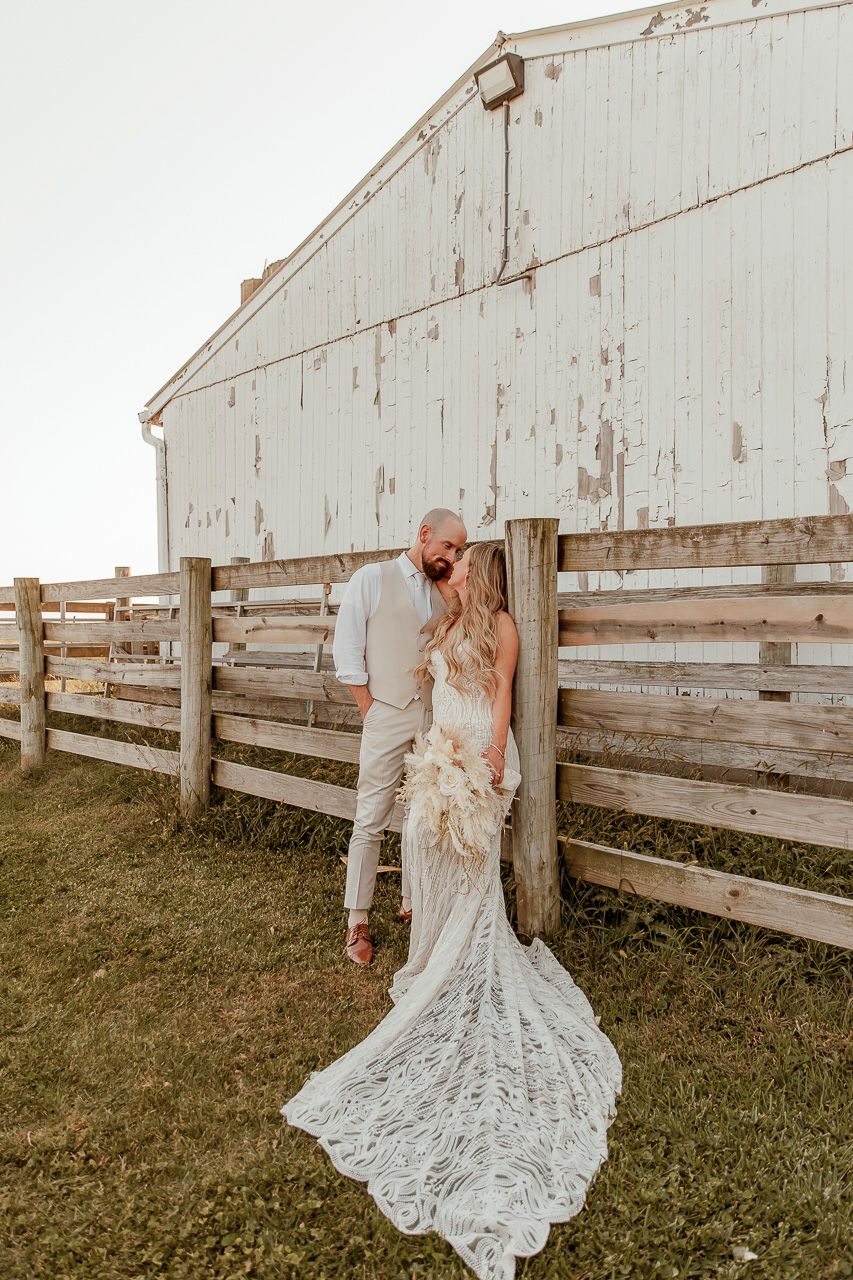 Bride in flowing lace gown poses with partner by weathered white barn and wooden fence during sunset.