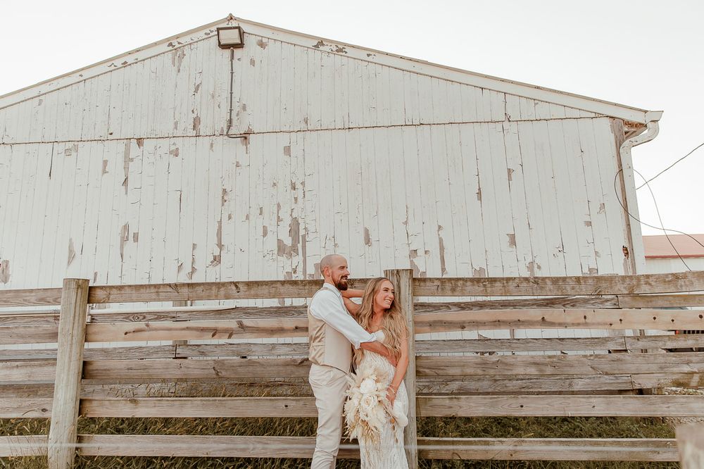 A couple stands together against a rustic white barn and wooden fence at a countryside wedding venue.