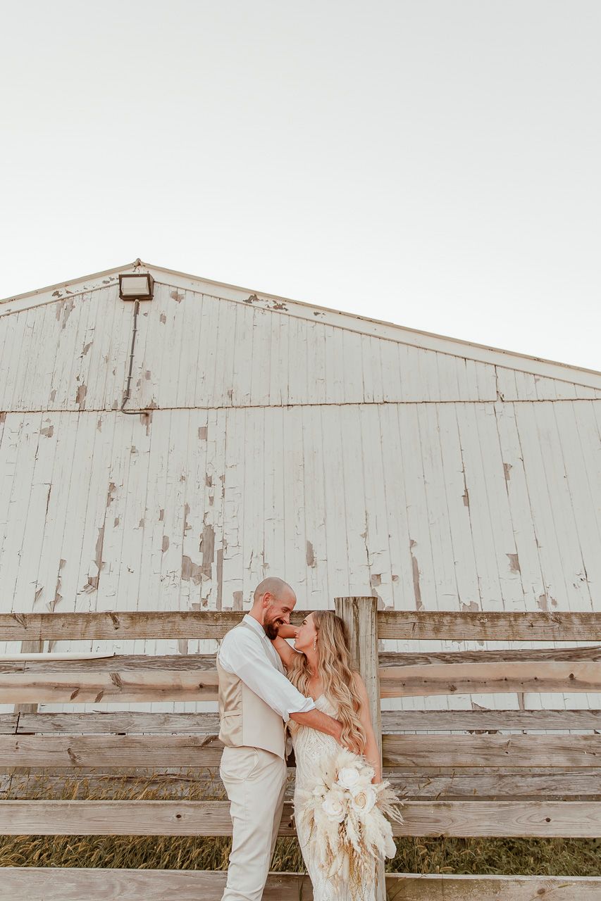 Couple embracing by weathered white barn wall during outdoor countryside photoshoot.