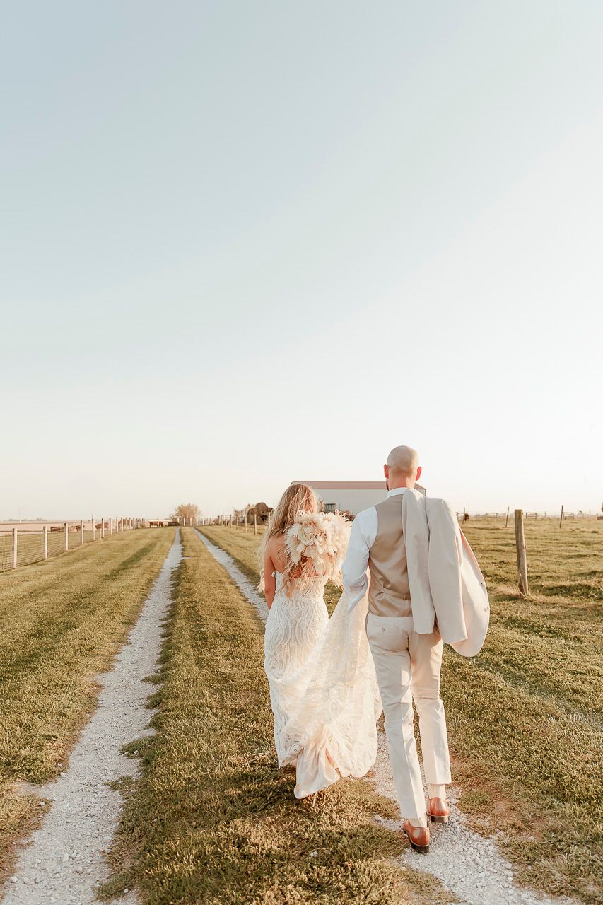 A couple in white attire walking down a rural path at sunset with a silo in the background.