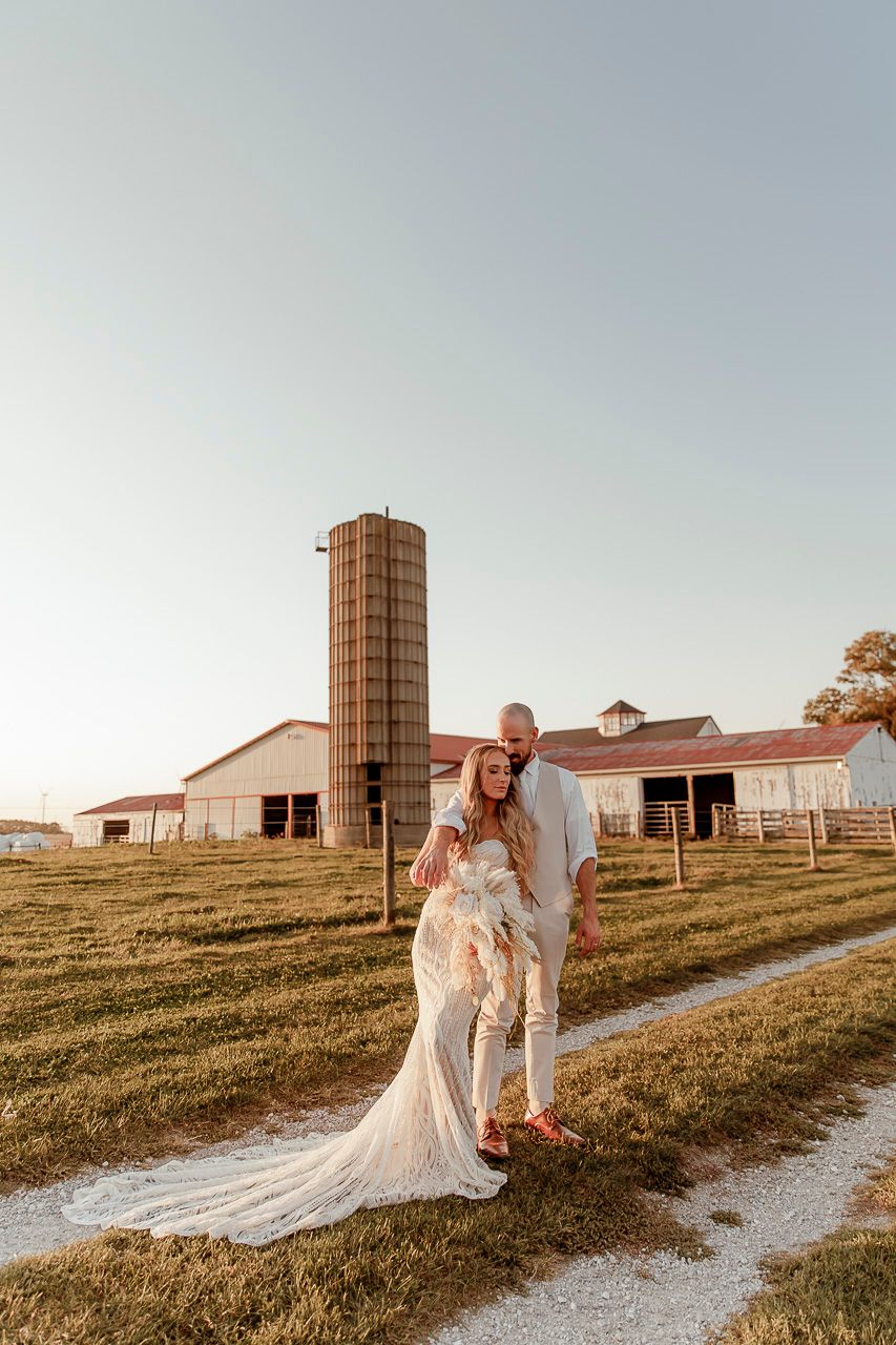 A couple poses together in front of a rustic barn and silo during golden hour on a scenic farm path.