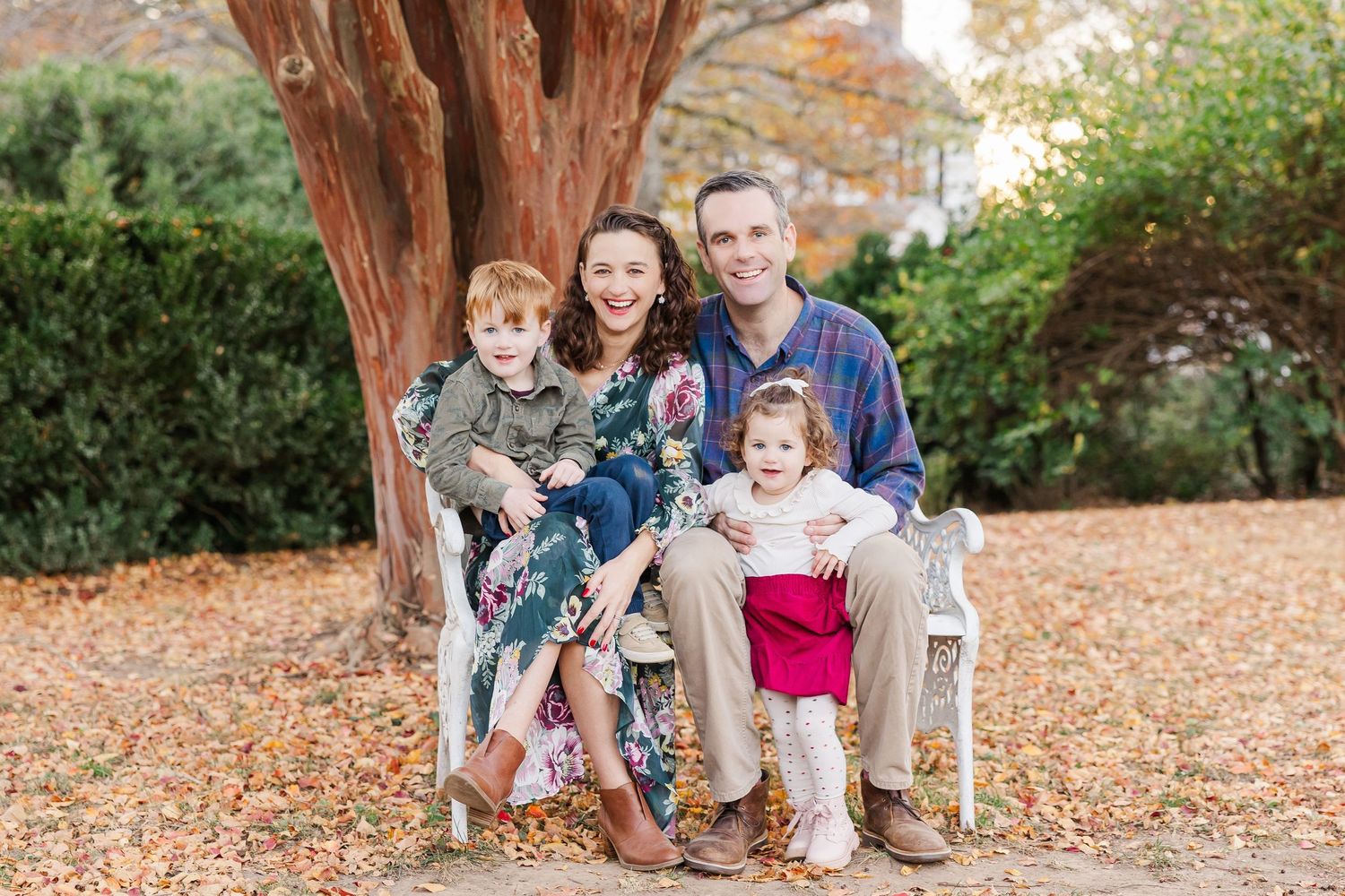 A family sits together on a wooden chair under a large tree with autumn leaves scattered on the ground.