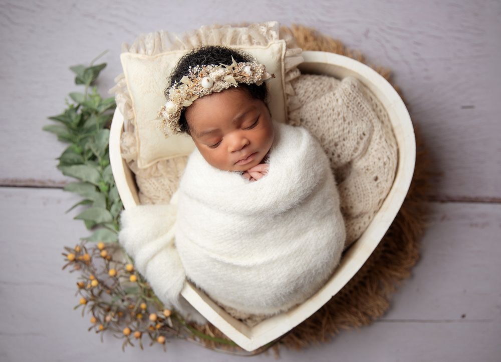 Sleeping newborn wrapped in white swaddle blanket rests in heart-shaped basket with floral accents and delicate headpiece.