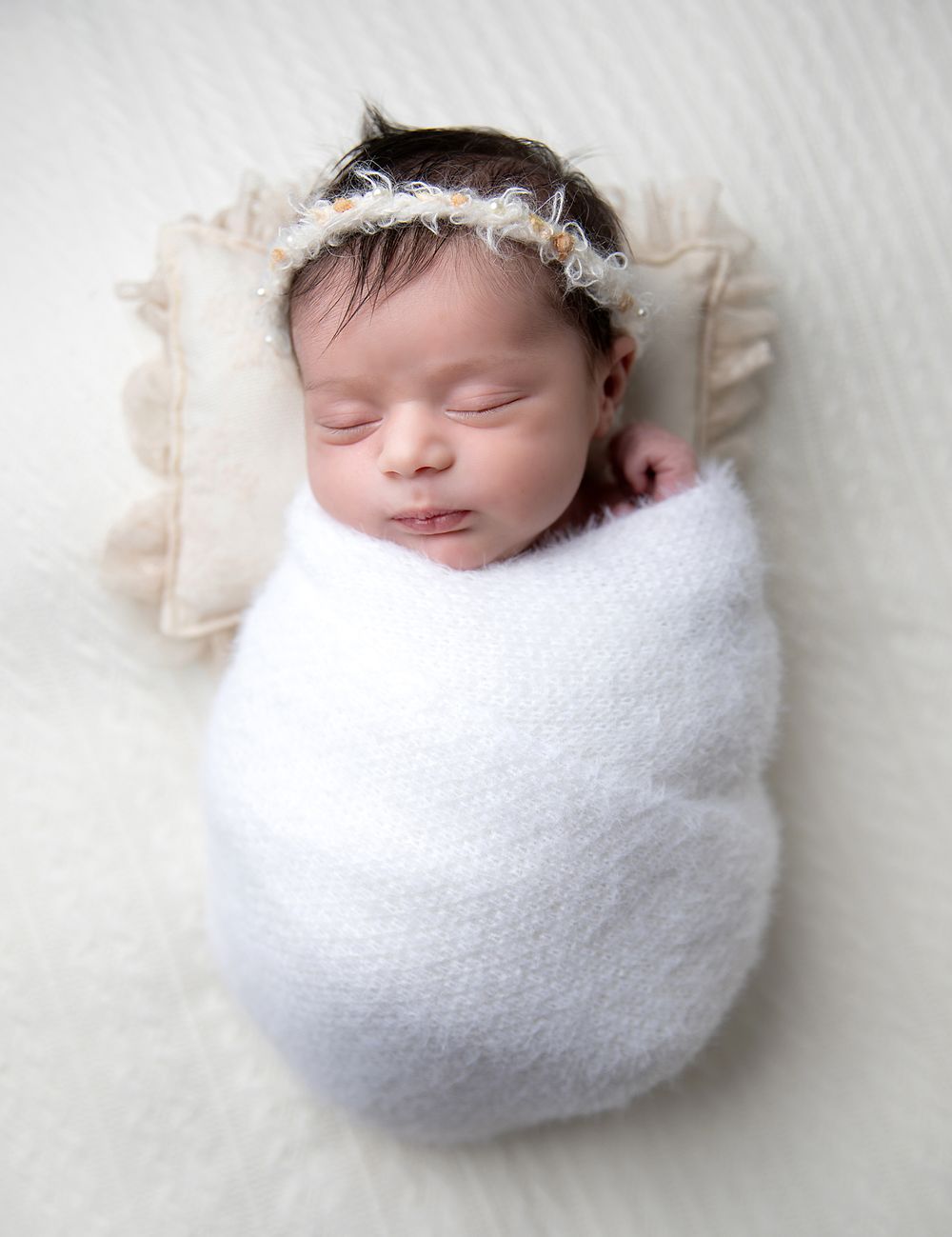 Peaceful sleeping newborn wrapped in soft white swaddle with delicate lace headband resting on cream pillow.
