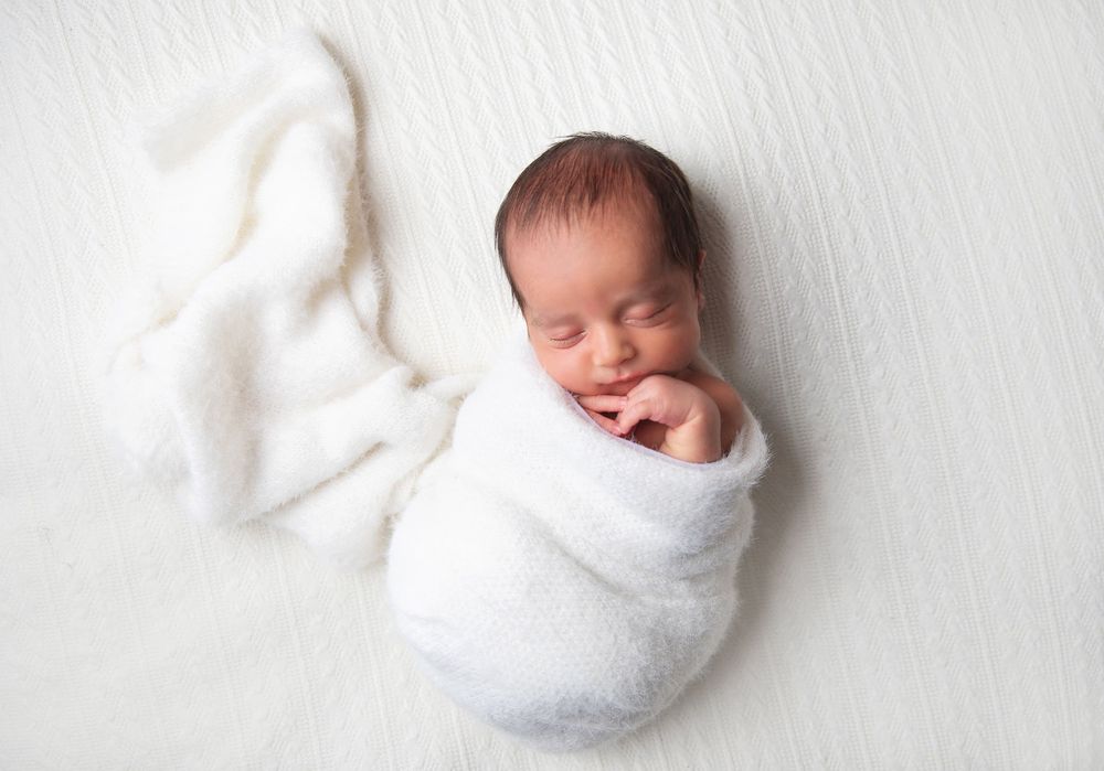 Sleeping newborn baby wrapped snugly in white swaddle blanket against white background.