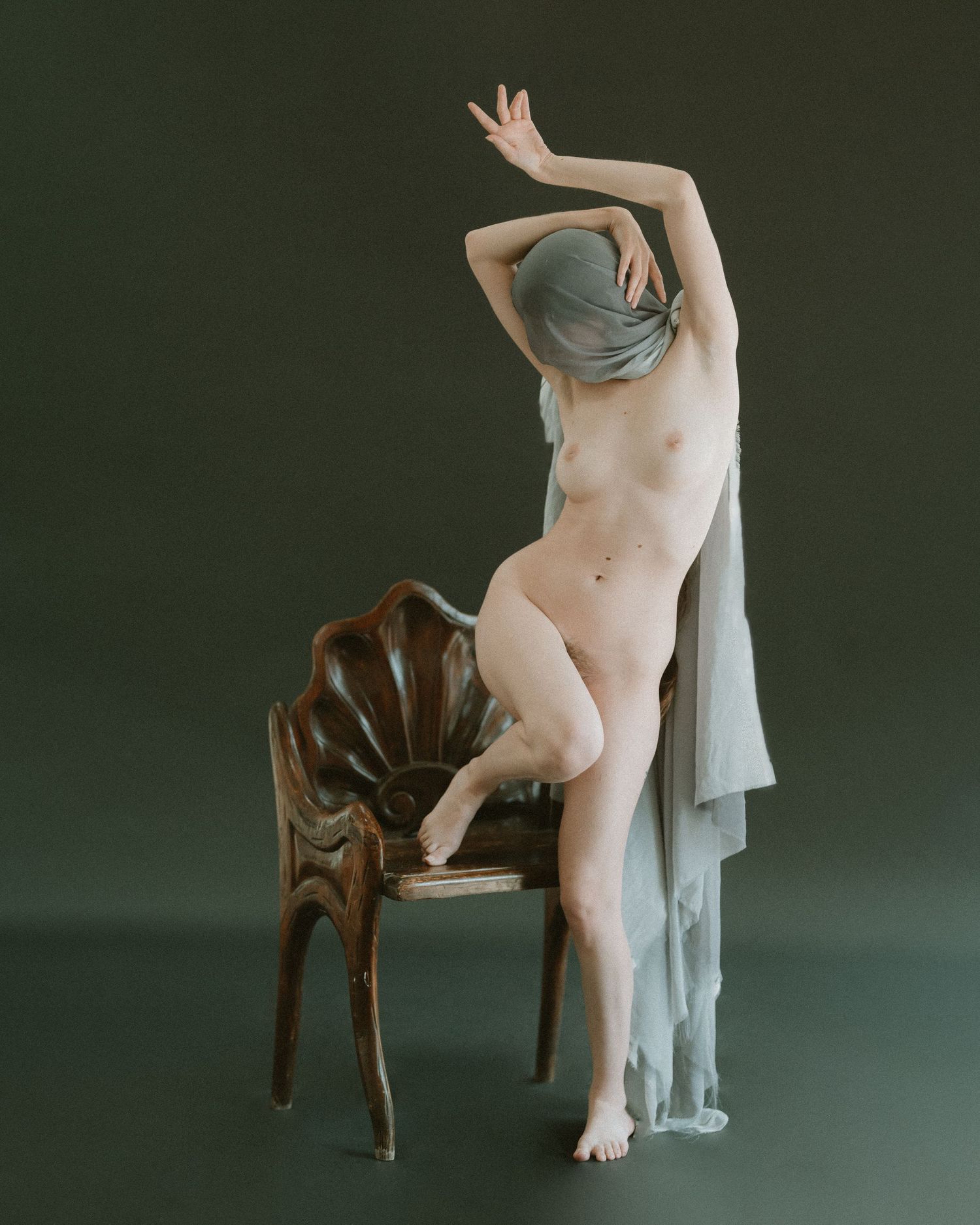 Graceful nude pose incorporating wooden chair and sheer fabric in studio setting.