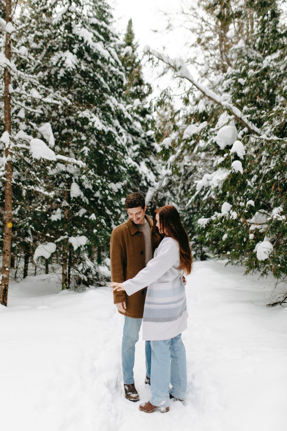 A couple shares an intimate moment while standing in a snow-covered forest with evergreen trees in winter.