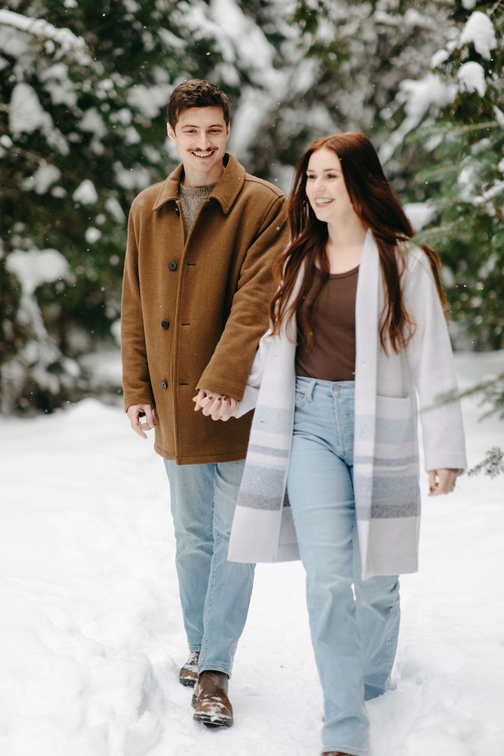 A couple walks hand in hand through a snowy forest path, wearing winter coats and coordinated casual outfits.