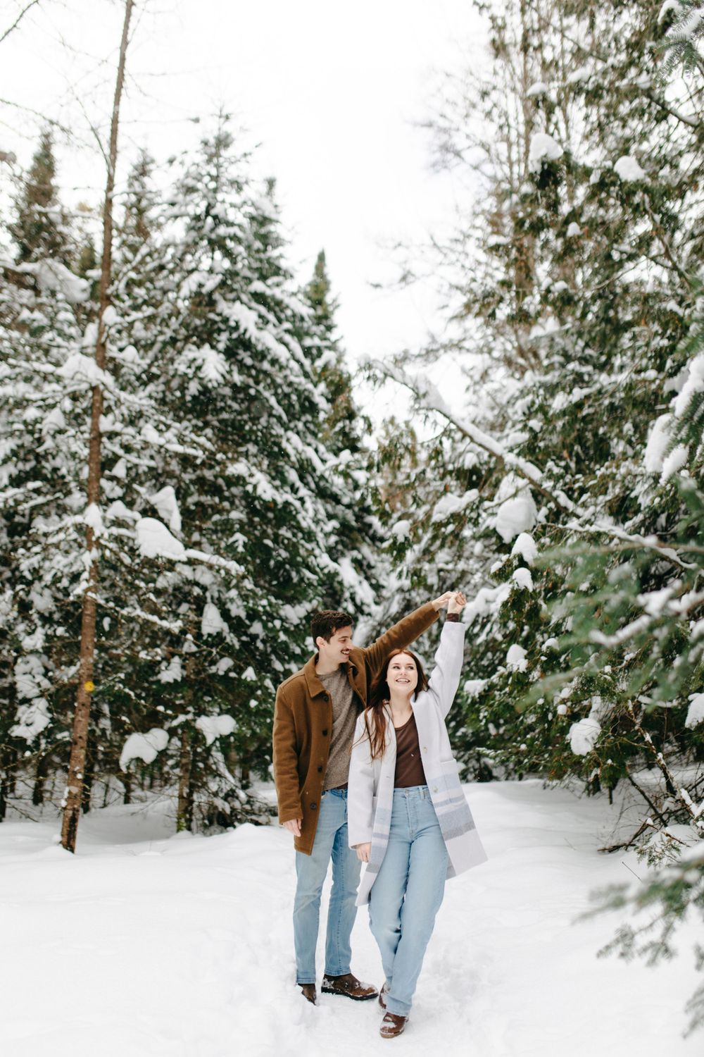 Two people walk through a snowy winter forest with snow-covered evergreen trees surrounding them.