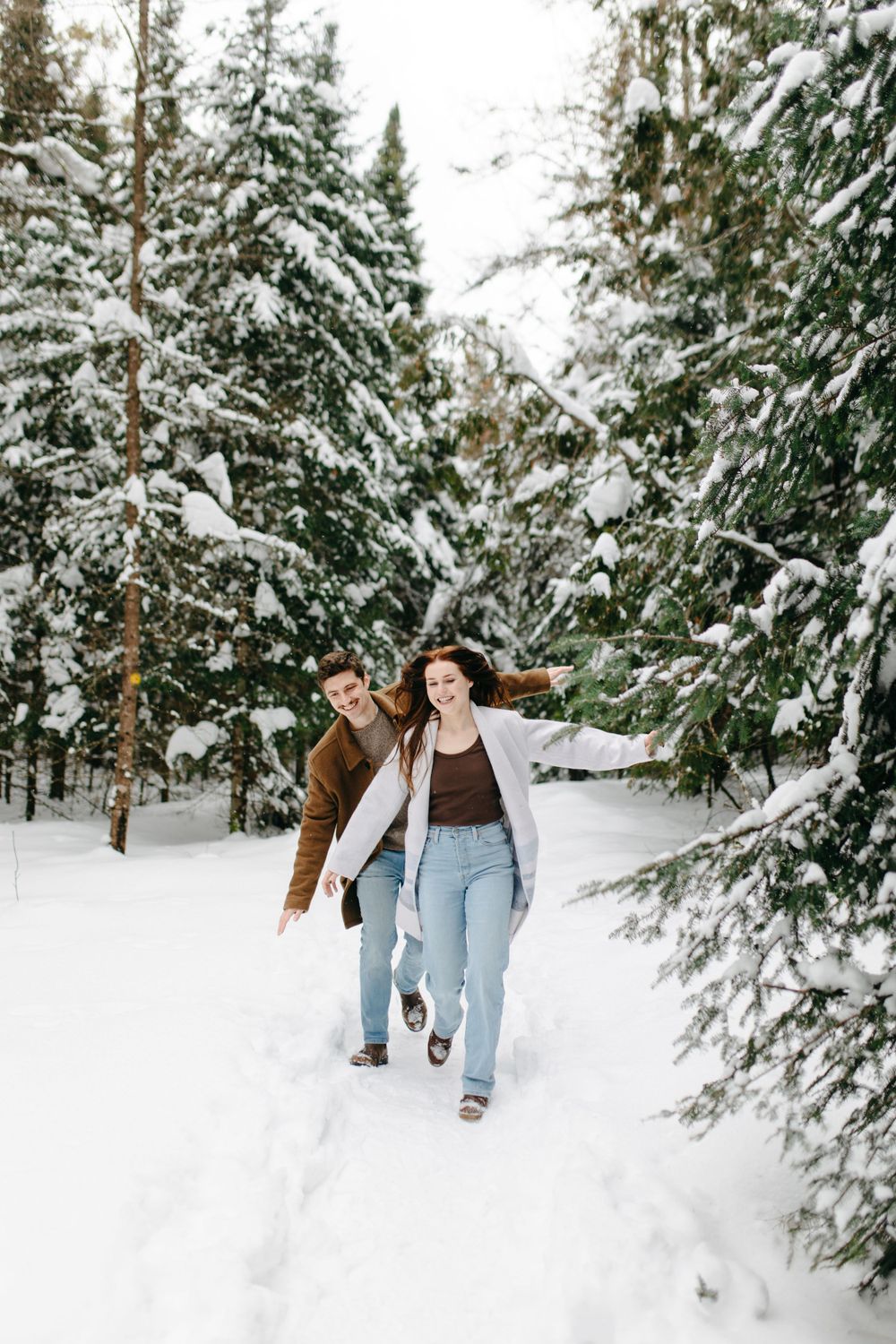 A couple walks hand in hand through a snow-covered forest trail surrounded by evergreen trees laden with fresh snow.