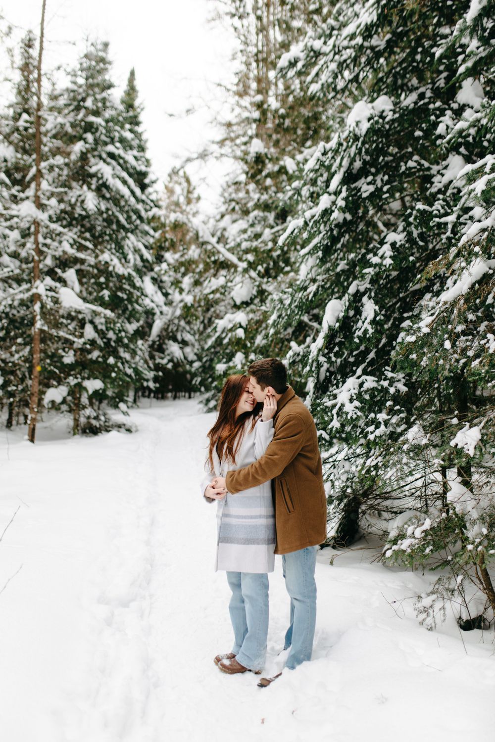A couple embraces on a snowy forest path surrounded by snow-covered evergreen trees in winter.