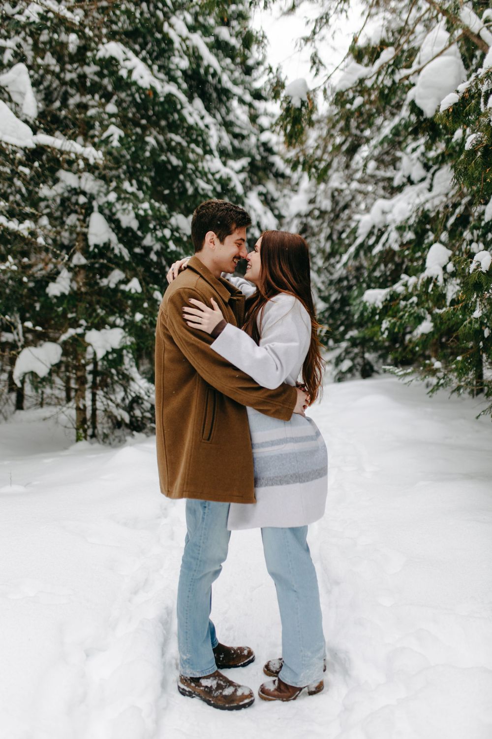 A couple embraces in a snowy forest setting wearing winter coats and matching boots amidst snow-covered evergreens.