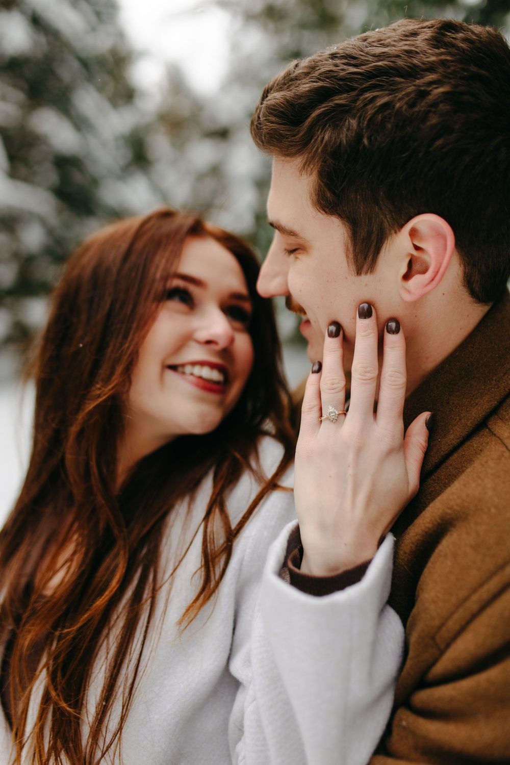 A couple shares an intimate winter engagement moment as she displays her ring while touching his face.